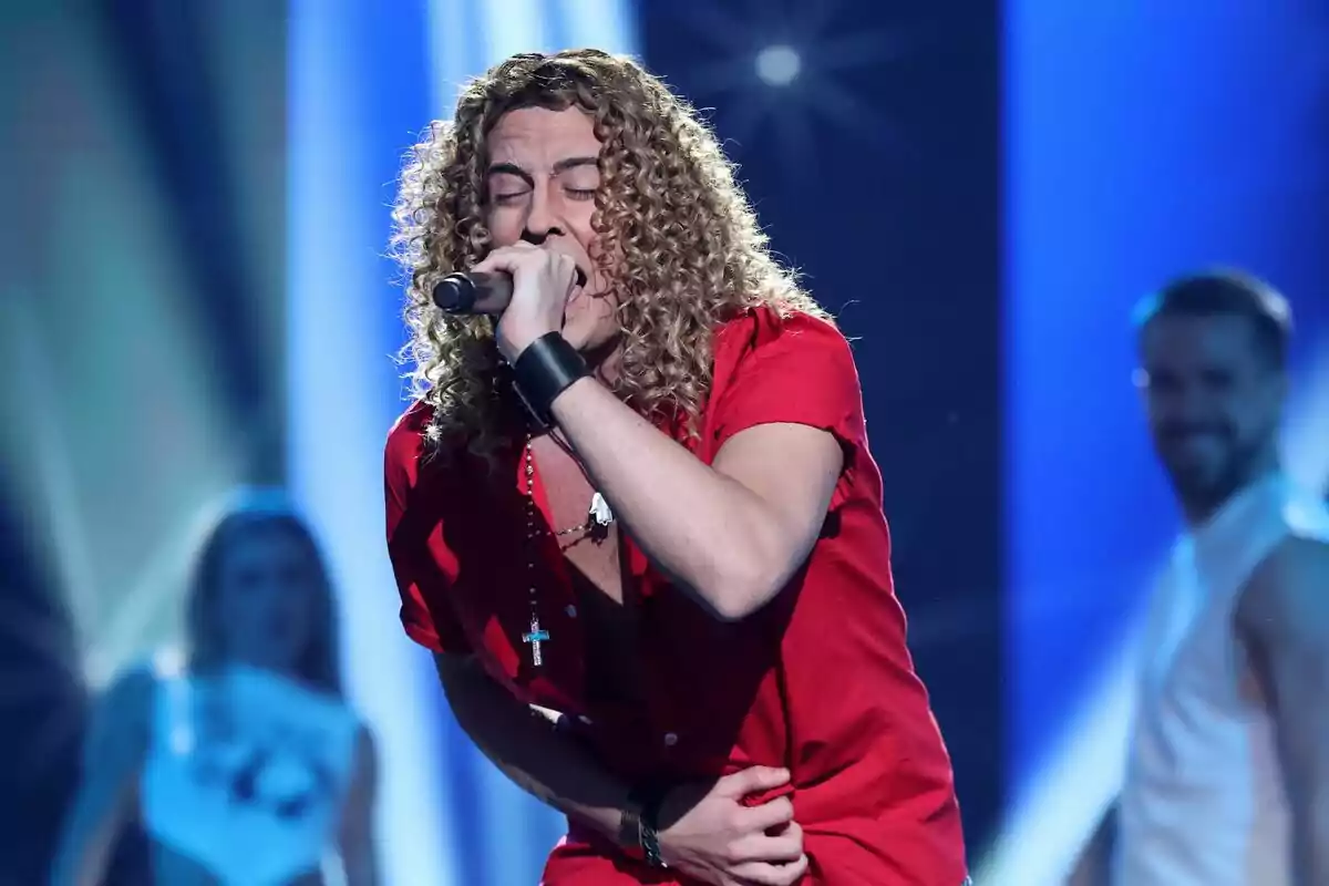 Mikel Hergoz con cabello rizado y camisa roja está actuando en el escenario mientras sostiene un micrófono en Tu Cara Me Suena. Mikel Hergoz con cabello rizado y camisa roja está actuando en el escenario mientras sostiene un micrófono en Tu Cara Me Suena.