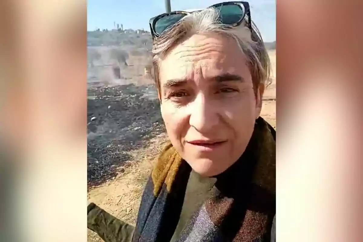 Una persona con gafas de sol en la cabeza está al aire libre en un área con vegetación quemada y humo en el fondo.