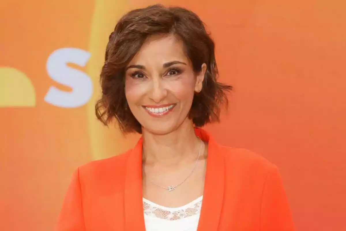 Adela González sonriente con cabello corto y ondulado, vestida con una chaqueta naranja, posando frente a un fondo naranja en Mañaneros