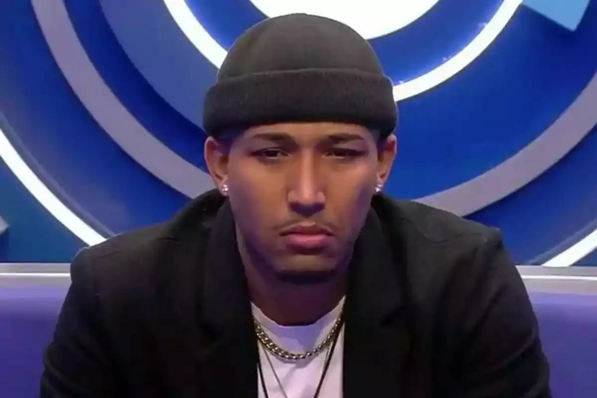 Adrián con gorro negro y chaqueta oscura está sentado frente a un fondo azul con círculos en Gran Hermano.