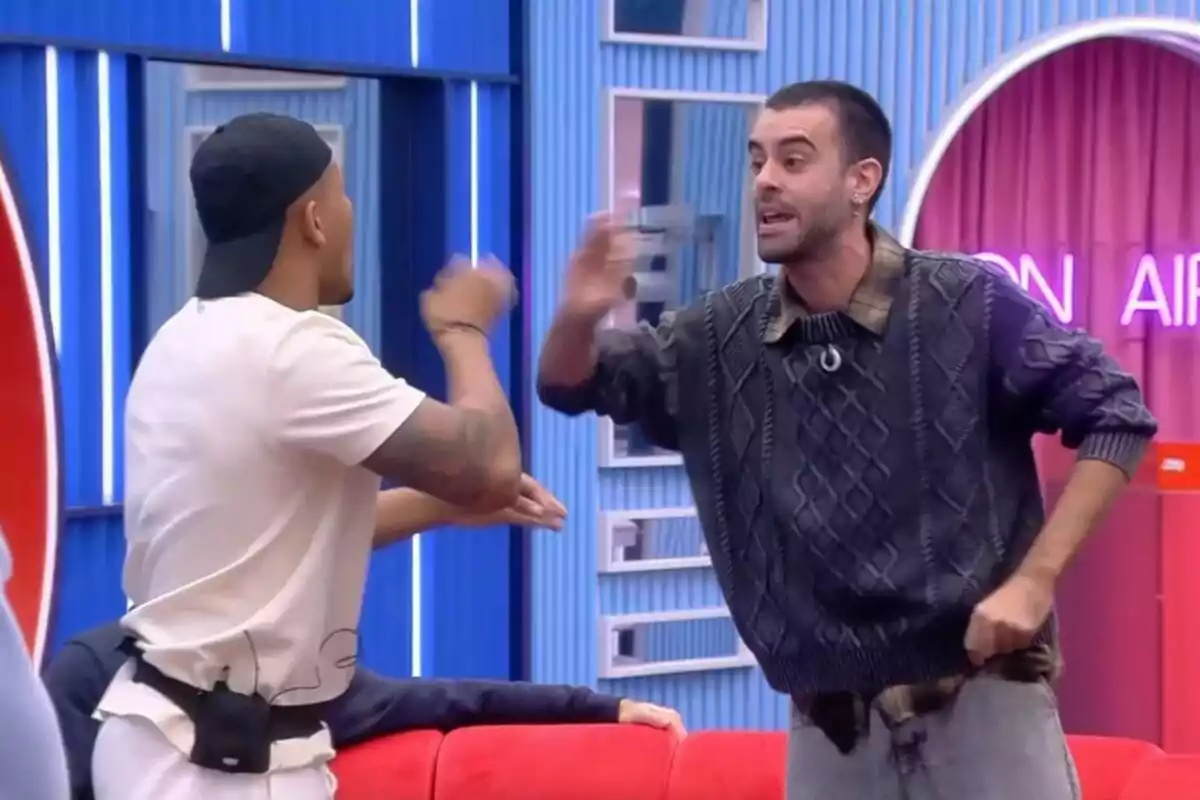 Adrián y Ruvens discutiendo en la casa de Gran Hermano con luces de neón y un sofá rojo.