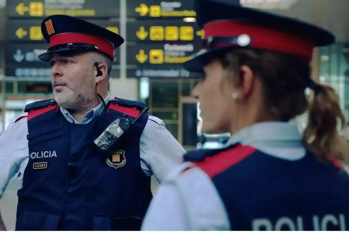 Dos agentes de policía con uniforme y gorra están de pie en un aeropuerto, uno de ellos lleva un dispositivo de comunicación en el chaleco.