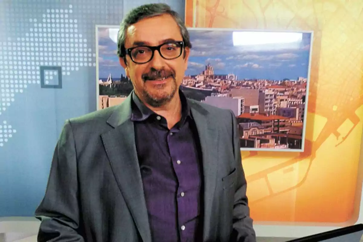 Agustí Forné con gafas y barba en un estudio de televisión con fondo de ciudad.