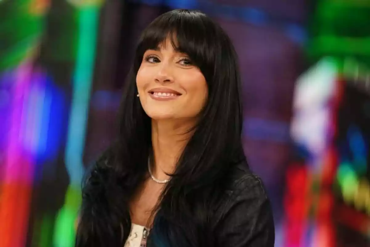 Aitana con cabello largo y oscuro con flequillo sonriendo frente a un fondo colorido y desenfocado en 'El Hormiguero'.