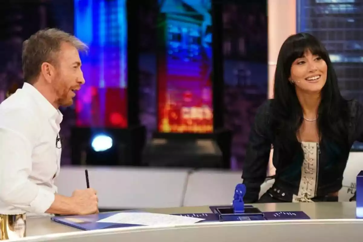 Aitana y Pablo Motos conversan y sonríen en 'La Revuelta' con luces de colores al fondo y papeles sobre la mesa. Aitana y Pablo Motos conversan y sonríen en 'La Revuelta' con luces de colores al fondo y papeles sobre la mesa.