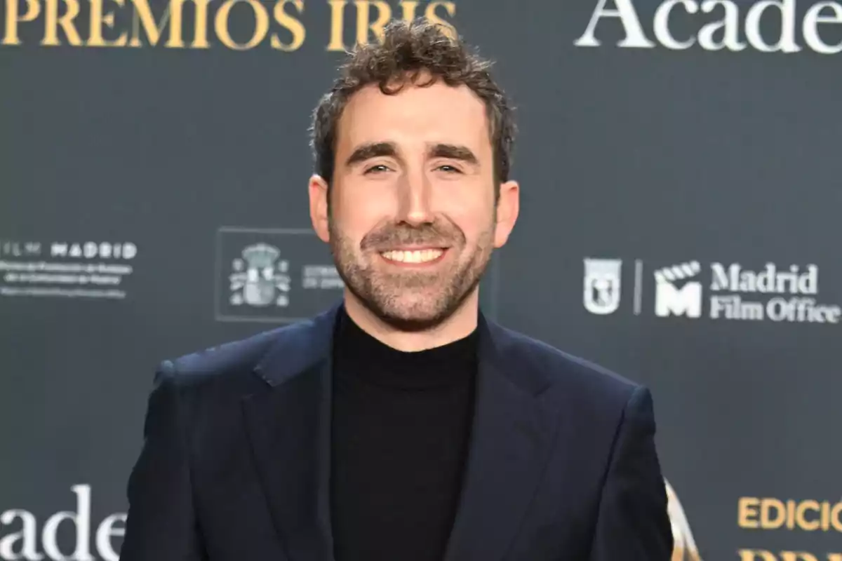 Aitor Albizua sonriente con barba y cabello rizado lleva un traje oscuro y está en un evento con un fondo que tiene logotipos y texto. Aitor Albizua sonriente con barba y cabello rizado lleva un traje oscuro y está en un evento con un fondo que tiene logotipos y texto.