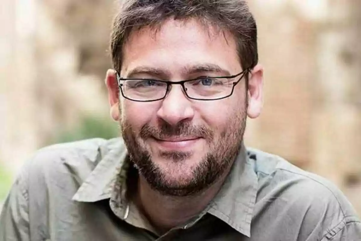 Un hombre con gafas y barba sonríe mientras mira a la cámara, vistiendo una camisa de color claro.