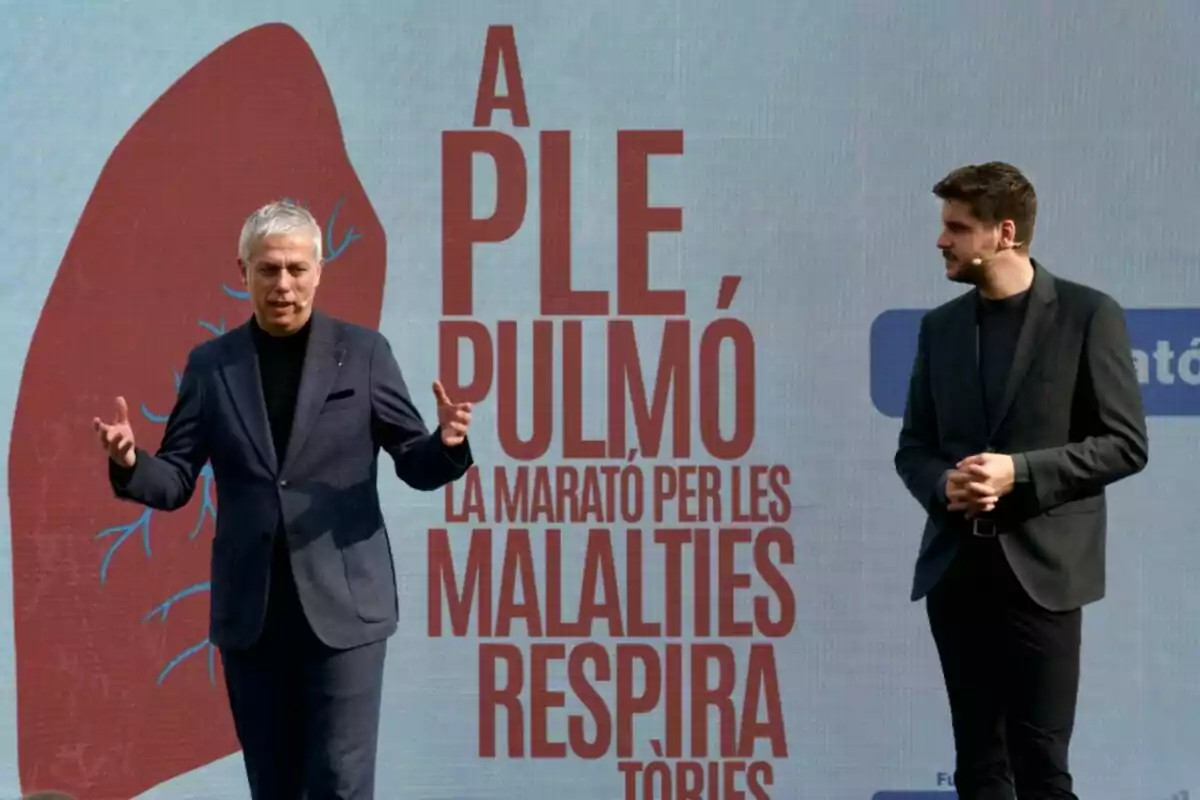 Dos personas de pie en un escenario al aire libre con un cartel de fondo que dice Dos personas de pie en un escenario al aire libre con un cartel de fondo que dice