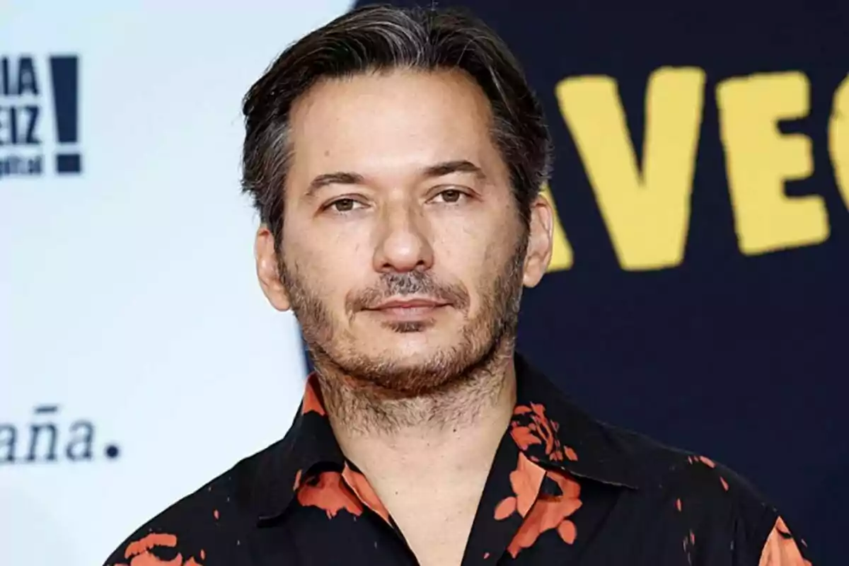 Alberto Caballero con barba y cabello oscuro lleva una camisa negra con detalles rojos frente a un photocall de La que se avecina.