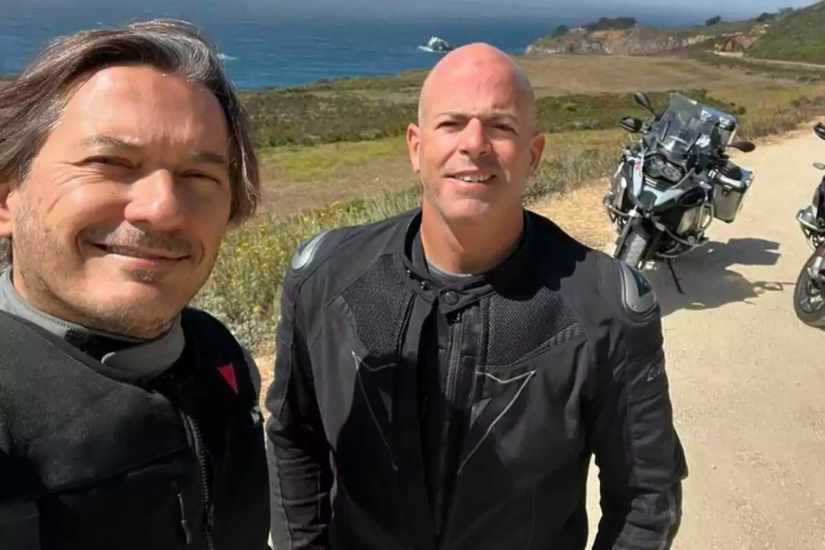 Alberto Caballero junto a Daniel Deorador con chaquetas de motociclista posan para una selfie al aire libre con motocicletas y un paisaje costero de fondo.