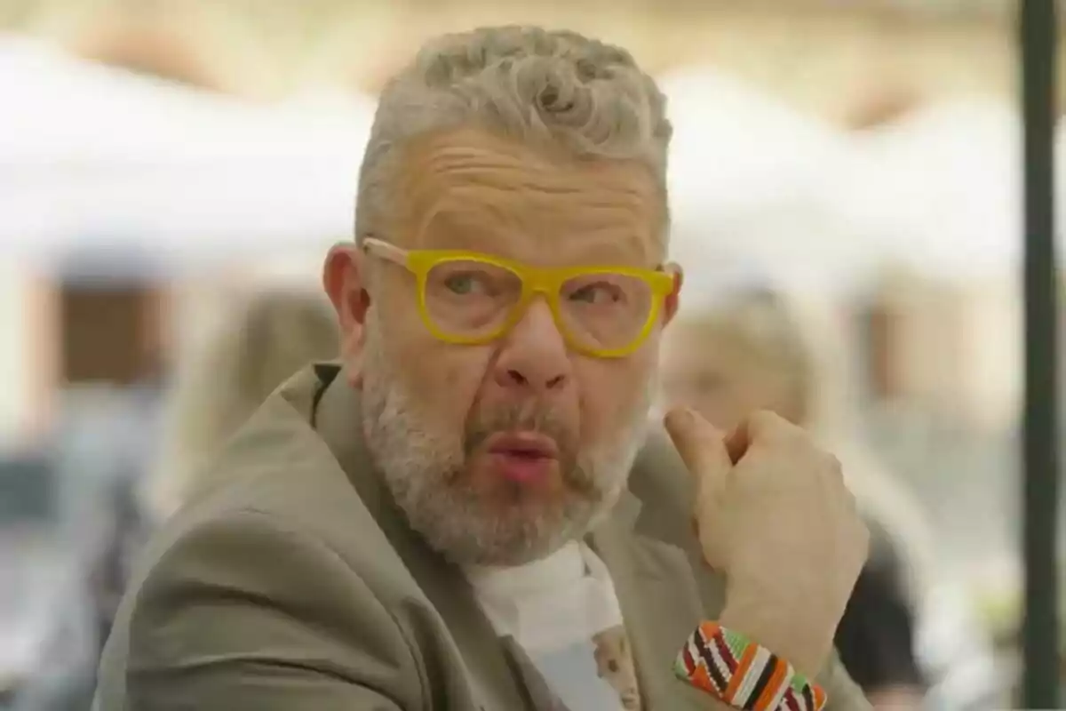 Alberto Chicote con gafas amarillas y barba canosa hace una expresión curiosa mientras está sentado al aire libre en Batalla de Restaurantes.