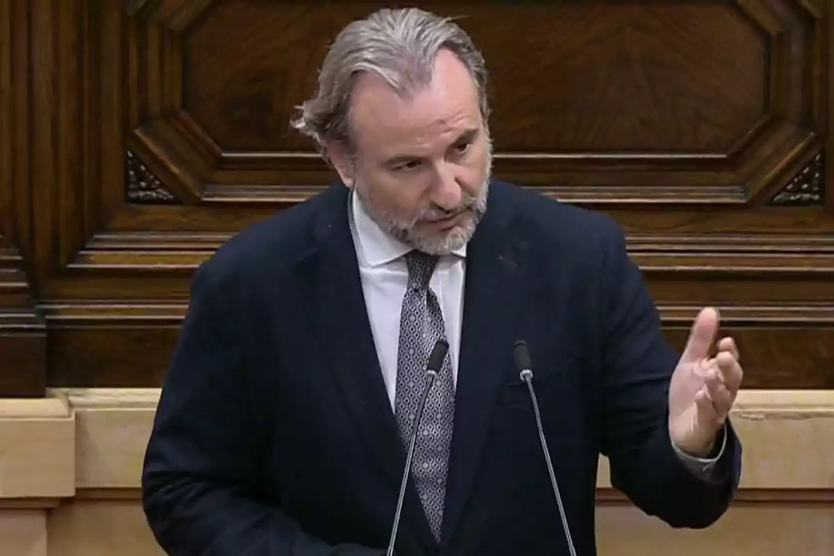 Un hombre de cabello canoso y barba está hablando en un podio con un fondo de madera decorativa.