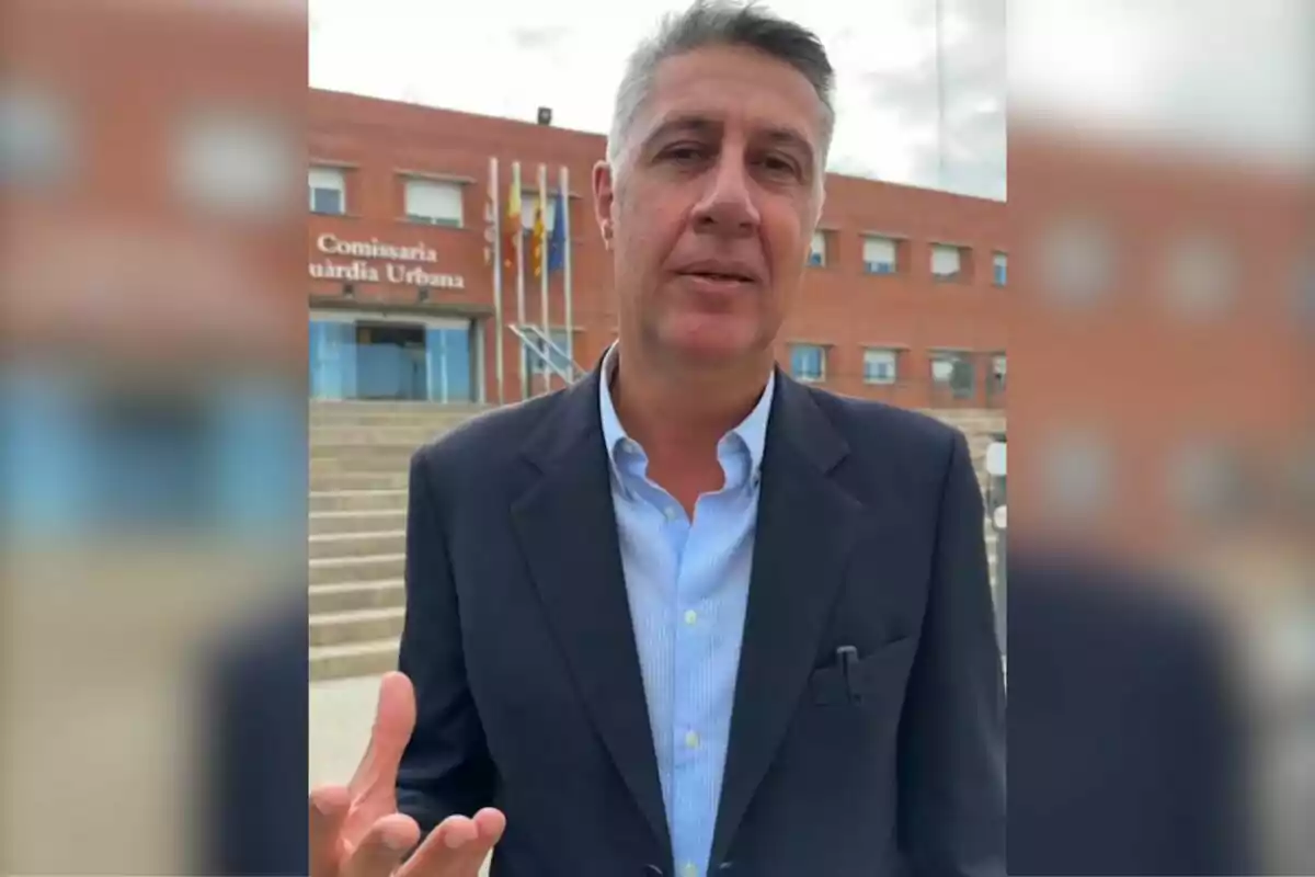 Un hombre de traje oscuro y camisa azul claro está de pie frente a un edificio de ladrillo con un letrero que dice "Comissaria Guardia Urbana" y varias banderas ondeando.