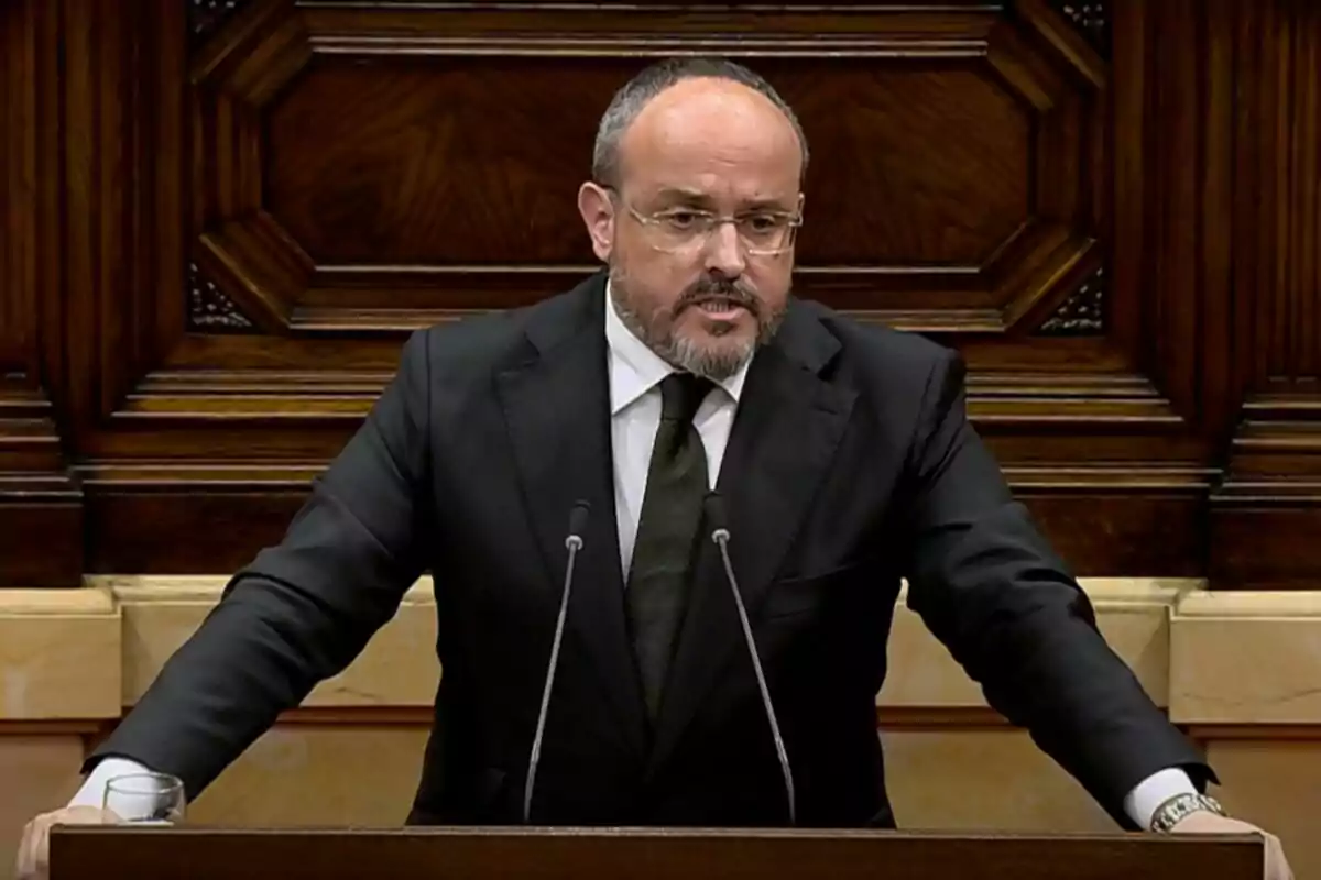 Un hombre con barba y gafas está hablando en un podio de madera con un fondo decorativo. Un hombre con barba y gafas está hablando en un podio de madera con un fondo decorativo.