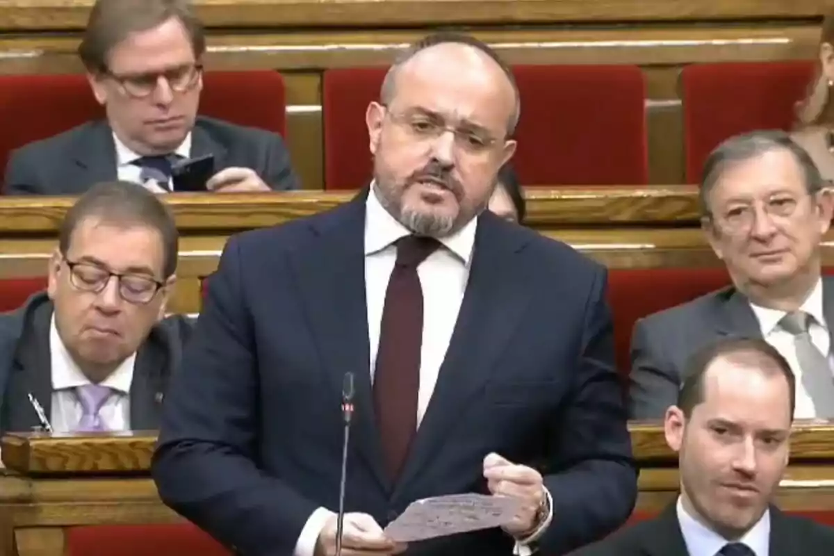 Un hombre de traje y corbata habla en un parlamento mientras otras personas lo observan desde sus asientos.