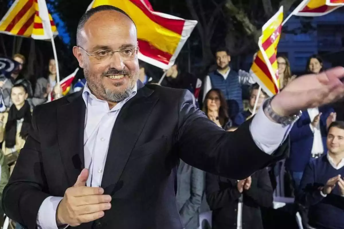 Alejandro Fernández en un mitin del PP sonriendo y alzando su mano izquierda con decenas de militantes detrás agitando banderas catalanas y españolas