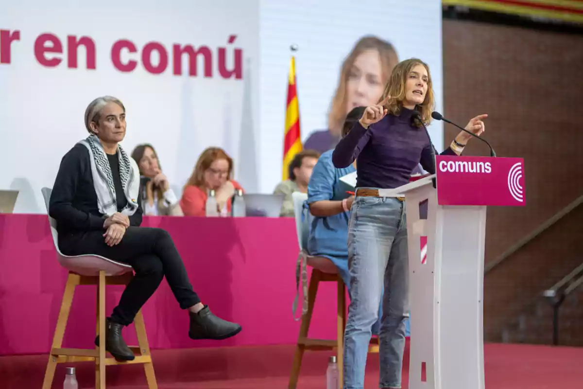 Jéssica Albiach hablando en un podio con el logo de "comuns" mientras Ada Colau está sentada cerca y varias personas están al fondo.