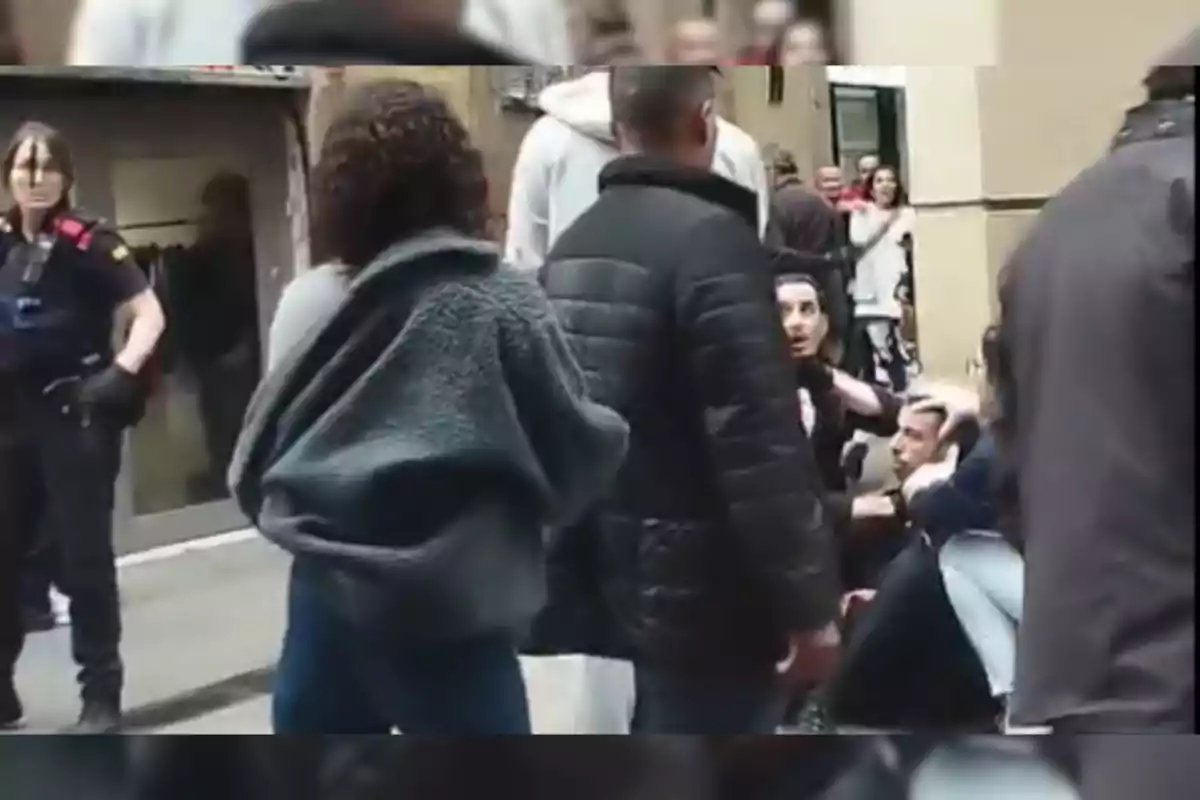 Un grupo de personas está reunido en una calle, algunas de pie y otras sentadas, mientras una oficial de policía observa la escena.