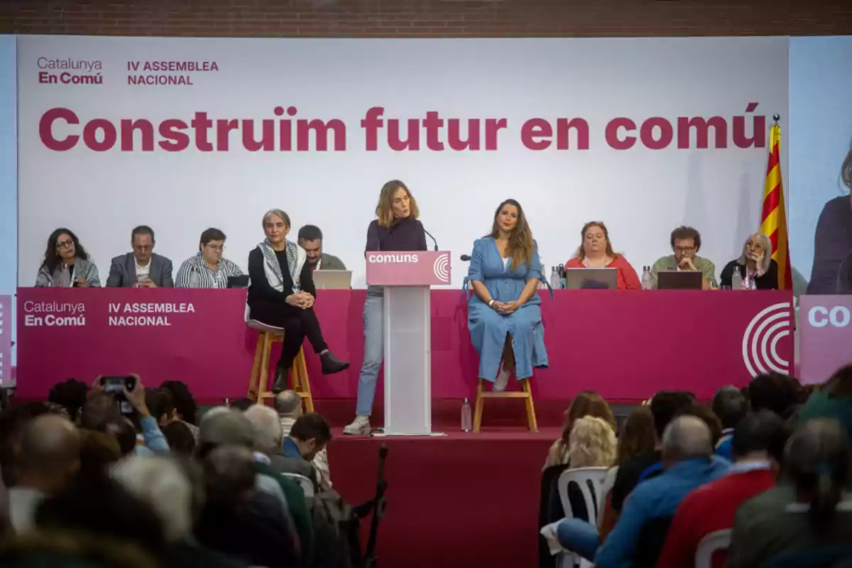 Una mujer habla en un podio durante una asamblea de Catalunya En Comú con varias personas sentadas detrás de ella y una audiencia al frente.