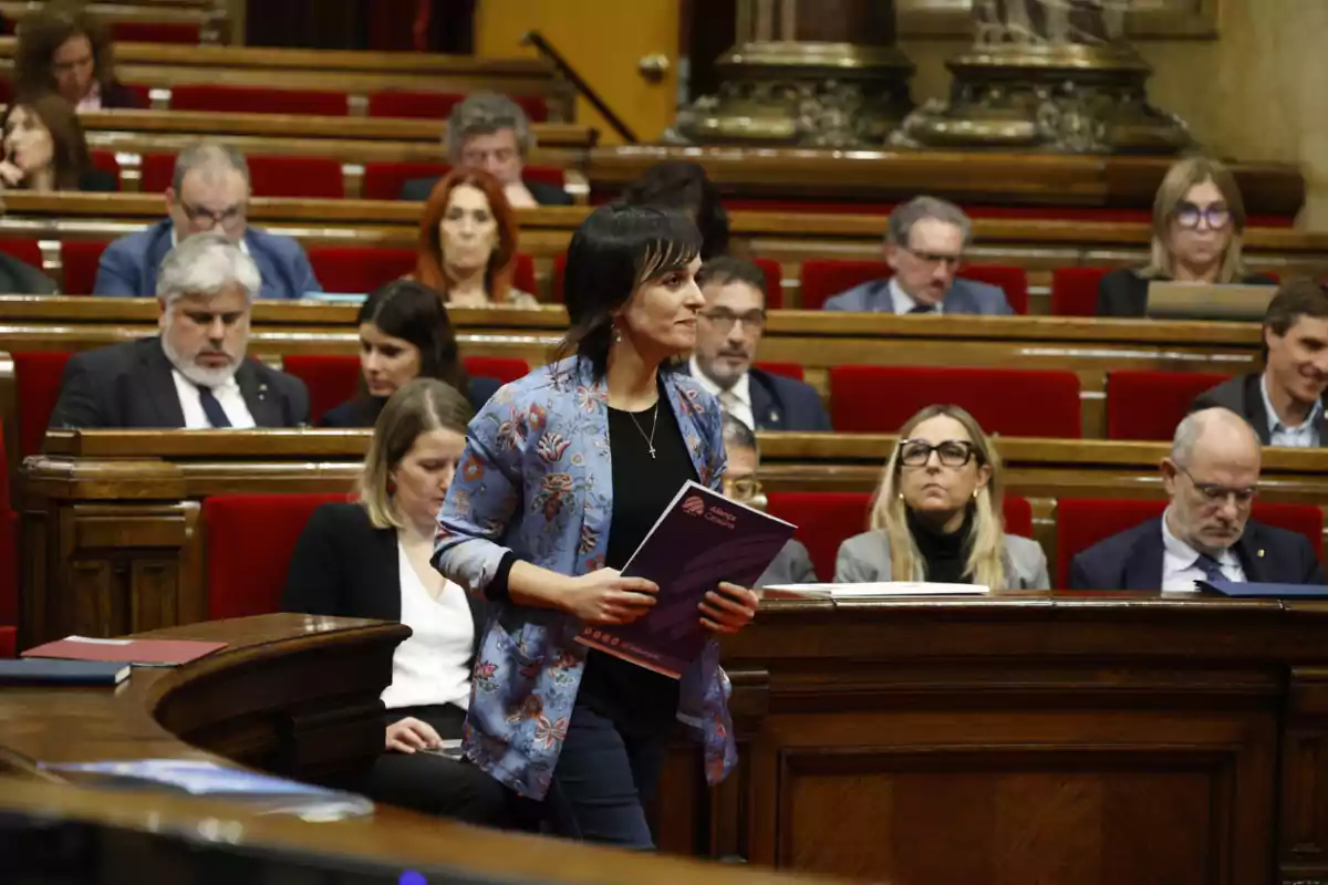 Una mujer de pie sosteniendo documentos en una sala de reuniones con varias personas sentadas en bancas rojas.