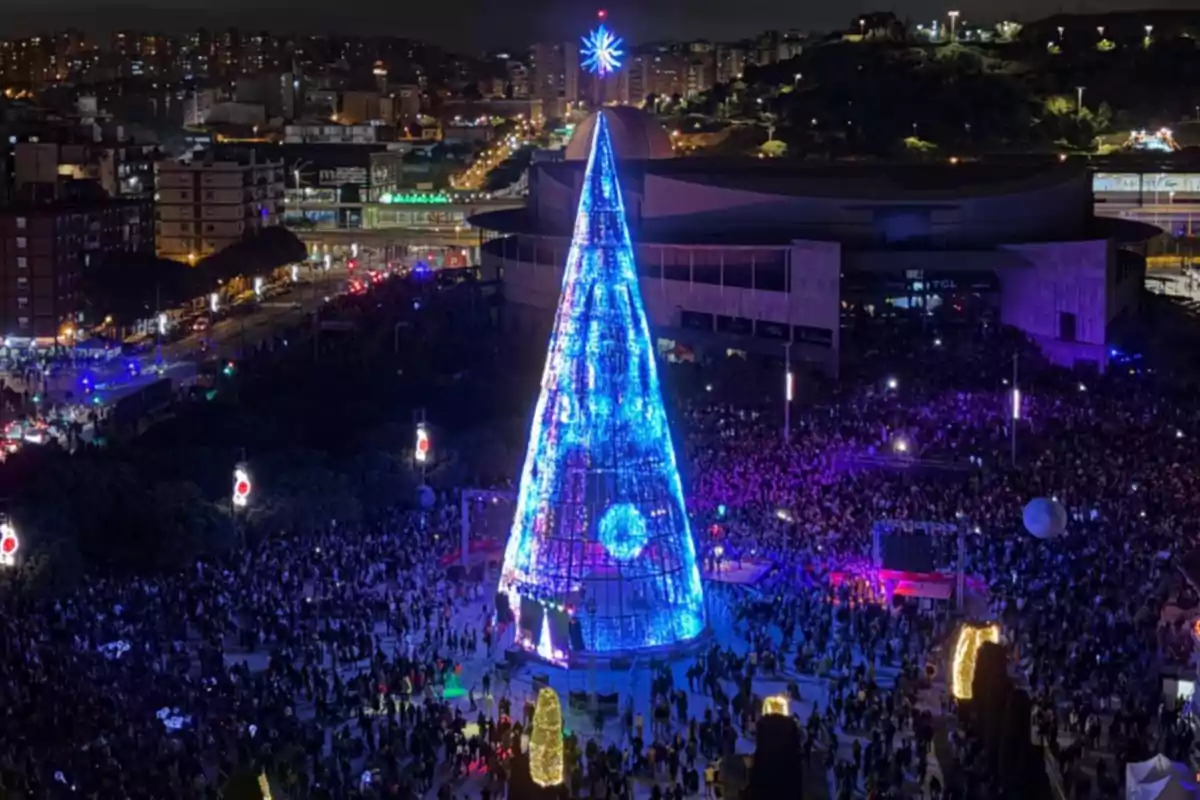 Una multitud se reúne alrededor de un gran árbol de Navidad iluminado en una plaza durante la noche con una ciudad de fondo.