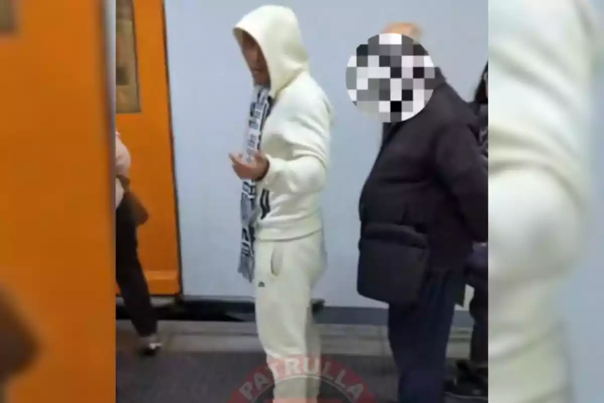 Dos personas de pie en una estación de metro una con sudadera blanca y capucha y la otra con el rostro pixelado.