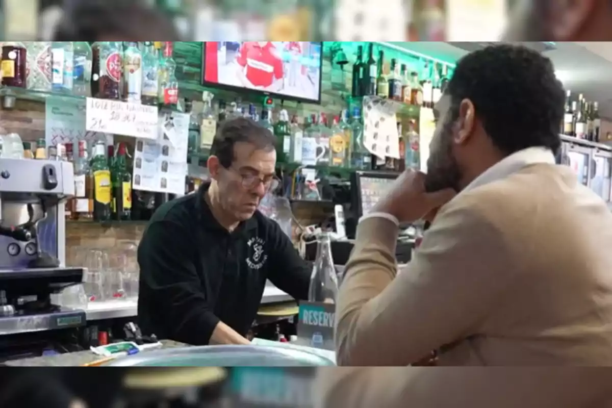 Un hombre detrás de la barra de un bar atiende a un cliente mientras prepara una bebida, rodeado de botellas y una pantalla de televisión en el fondo.