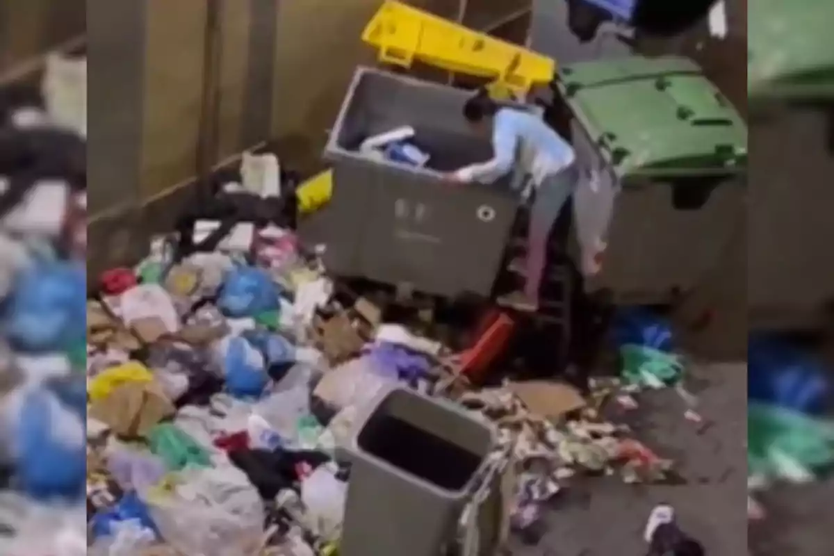 Una persona revisa el contenido de un contenedor de basura rodeado de bolsas y desechos esparcidos en el suelo.