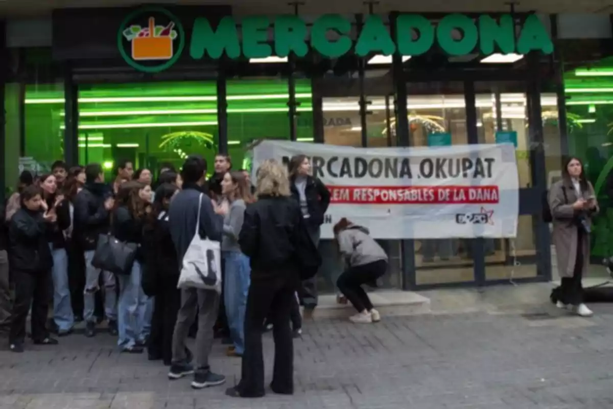 Un grupo de personas se reúne frente a la entrada de un supermercado Mercadona, donde hay una pancarta con un mensaje de protesta.