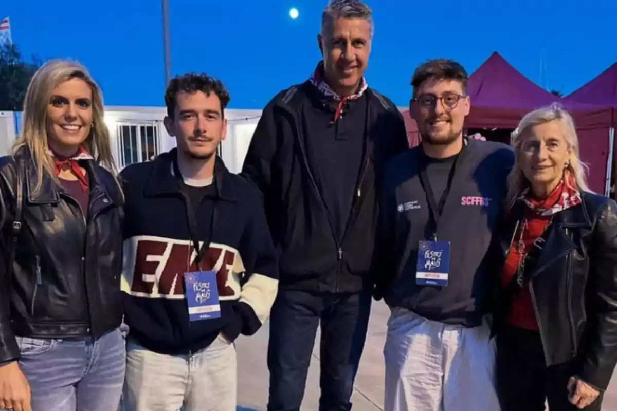Un grupo de cinco personas posa para una foto al aire libre durante un evento, con un cielo azul de fondo y carpas rojas detrás.