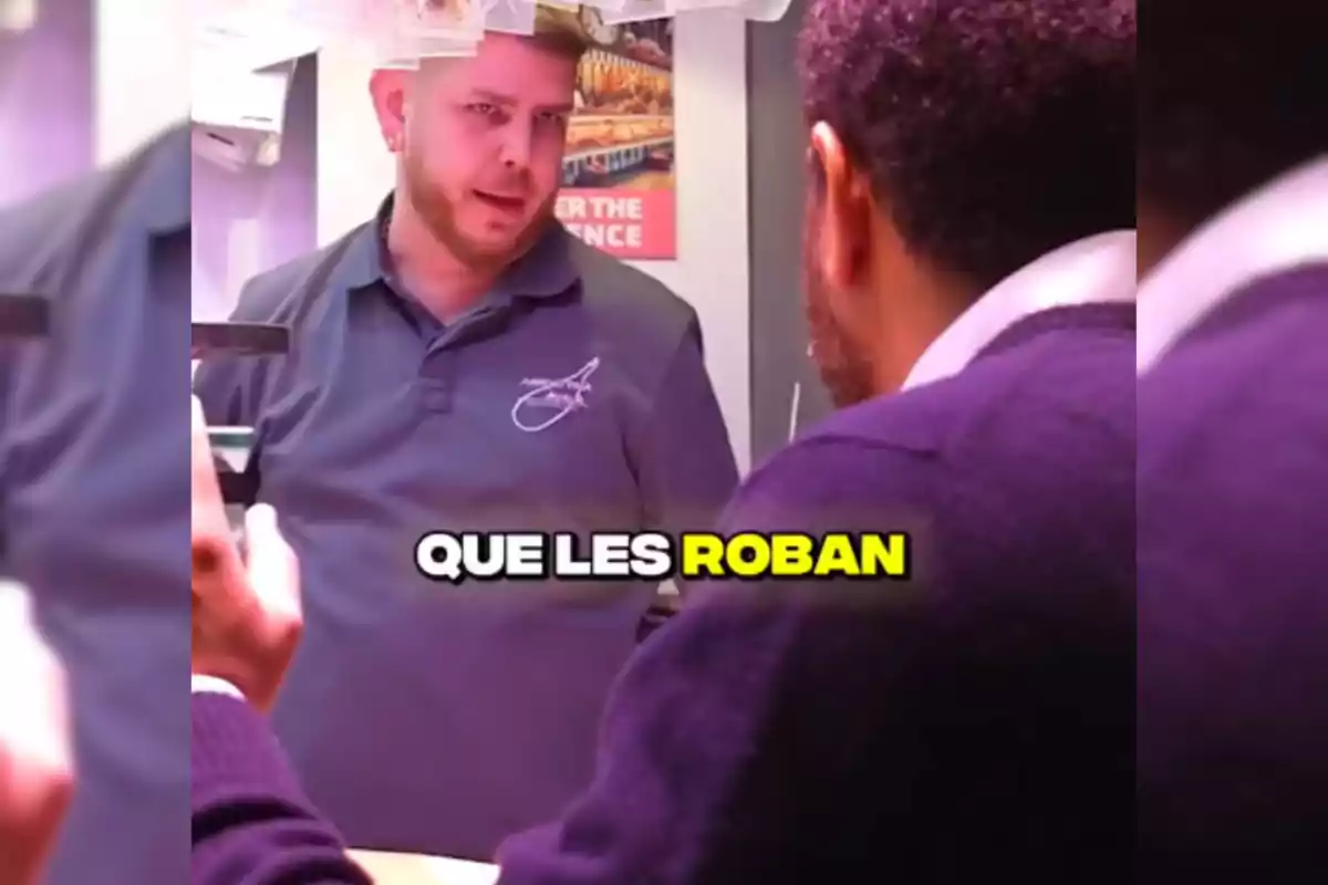 Dos personas conversan en un mostrador, una de ellas lleva una camiseta con un logotipo y la otra está de espaldas; en el centro de la imagen hay un texto que dice "QUE LES ROBAN".