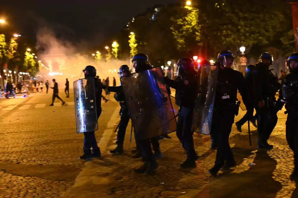 Policías antidisturbios con escudos avanzan por una calle iluminada de noche mientras al fondo se observa humo y personas dispersas