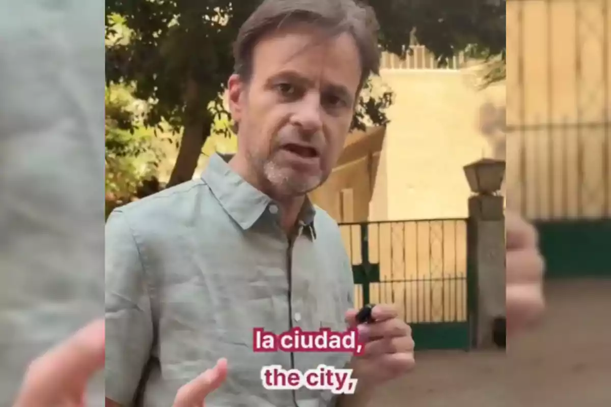 Hombre de cabello castaño y barba corta con camisa gris claro hablando al aire libre frente a una reja verde y árboles, con subtítulos en español e inglés que dicen la ciudad the city