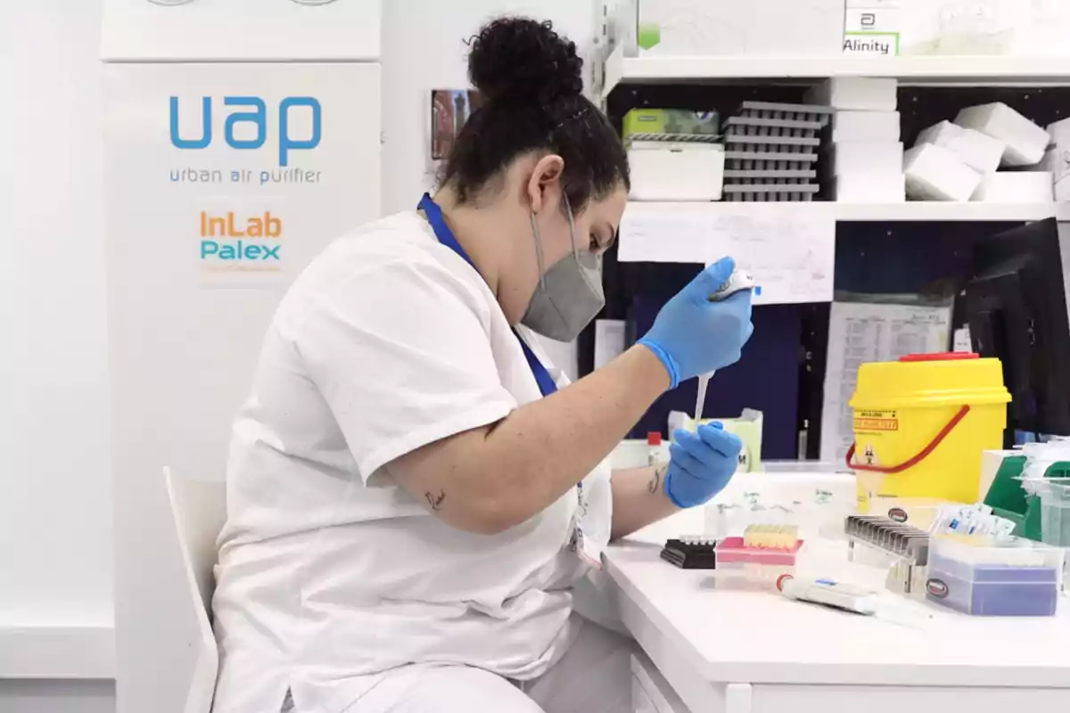 Una persona con bata blanca y guantes azules realiza una tarea de laboratorio, utilizando una pipeta en un entorno con varios equipos y suministros médicos.