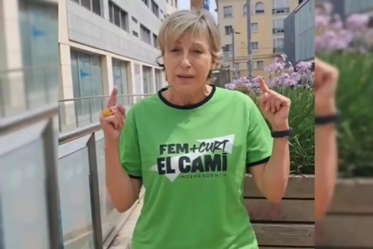 Carme Forcadell con una camiseta verde que dice "FEM + CURT EL CAMÍ INDEPENDÈNCIA" está levantando ambos dedos índices en un entorno urbano.