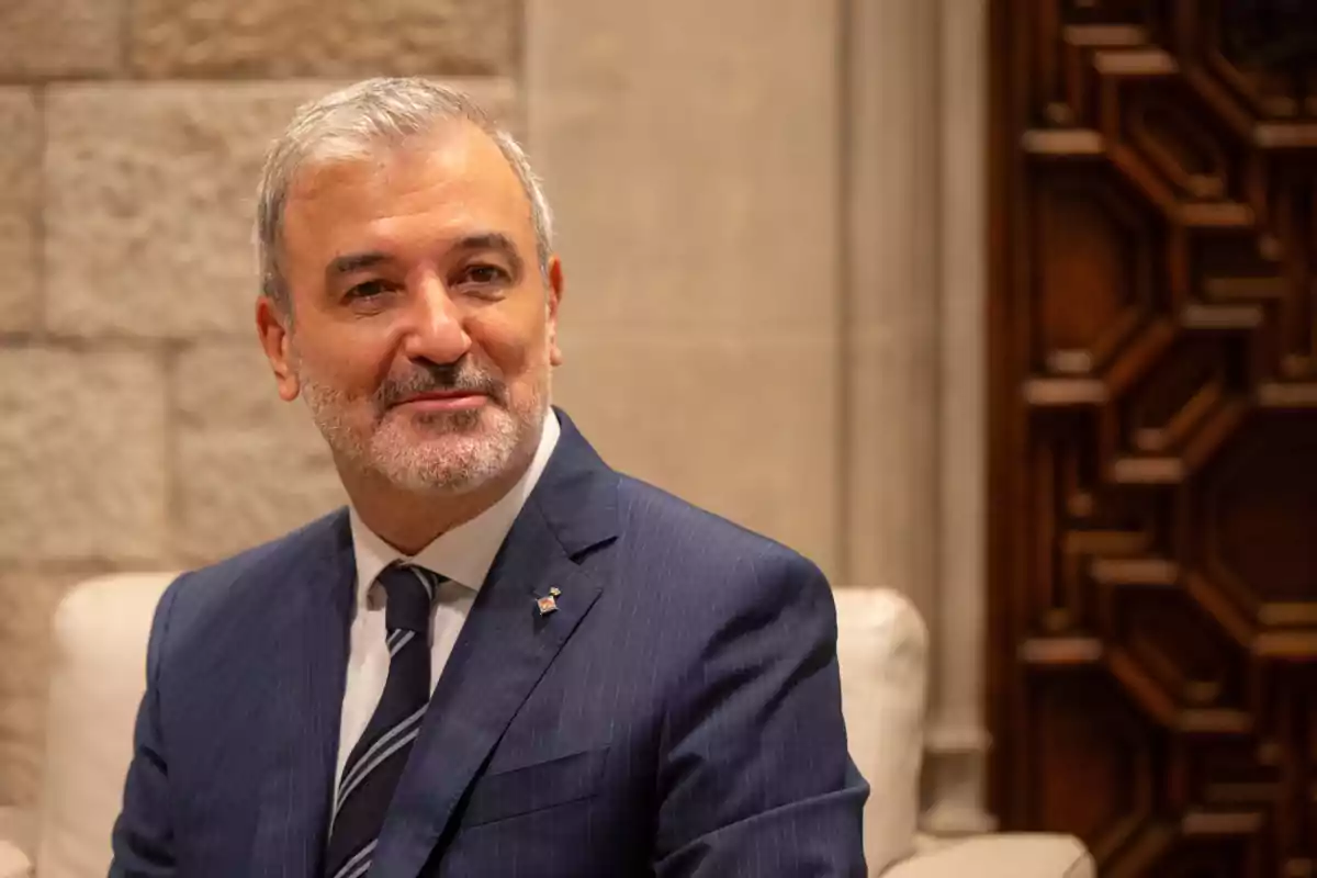 Jaume Collboni, el alcalde de Barcelona, vestido con un traje azul y corbata, está sentado en un entorno interior con paredes de piedra y una puerta de madera decorativa al fondo.