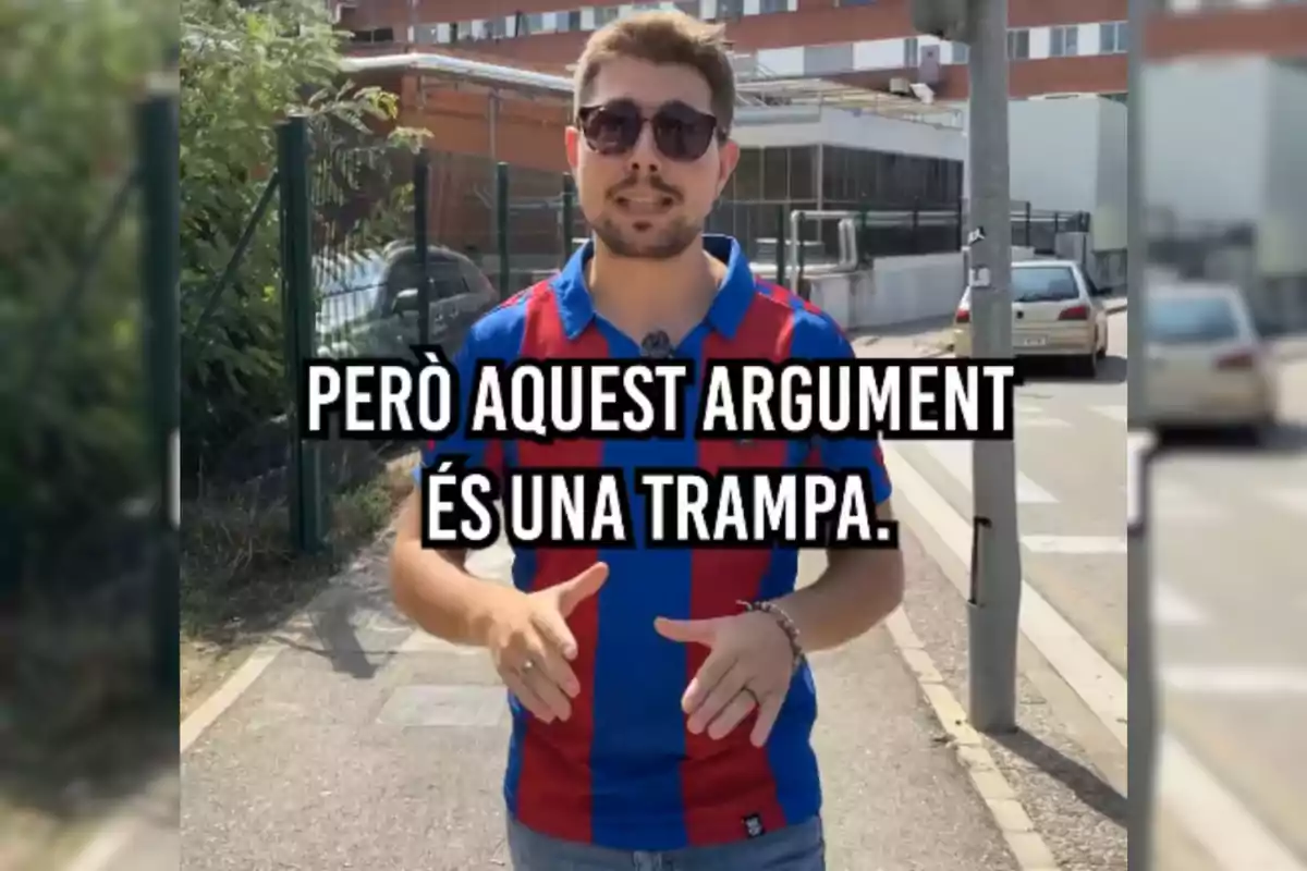 Un hombre de la CUP con gafas de sol y una camiseta de rayas rojas y azules está de pie en una acera, con un texto en catalán que dice "PERÒ AQUEST ARGUMENT ÉS UNA TRAMPA."