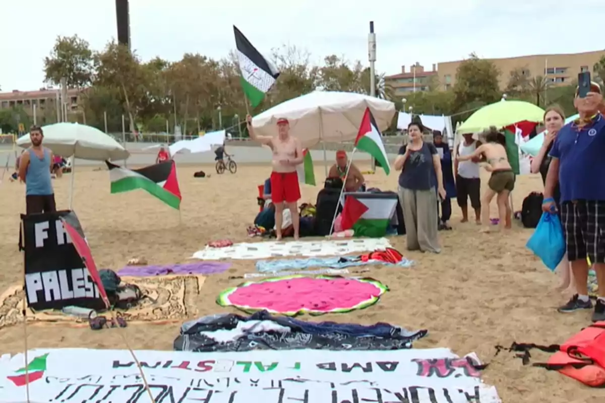 Personas en una playa con banderas palestinas y pancartas, algunas de las cuales están sentadas bajo sombrillas mientras otras están de pie, una de ellas sosteniendo una bandera.