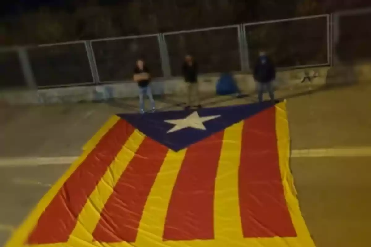 Tres personas de pie detrás de una gran bandera estelada en el suelo, con una valla metálica al fondo.