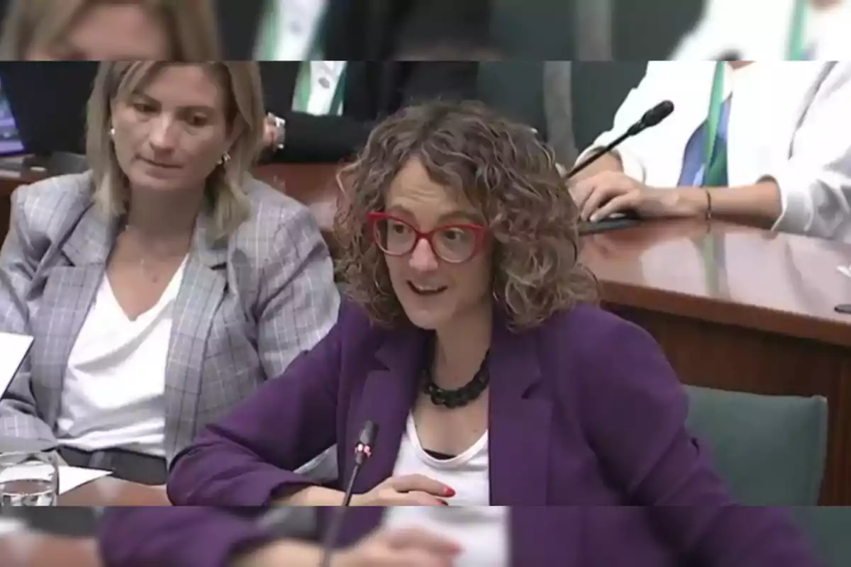 Dos mujeres sentadas en una mesa, una de ellas con gafas rojas y chaqueta morada hablando frente a un micrófono.