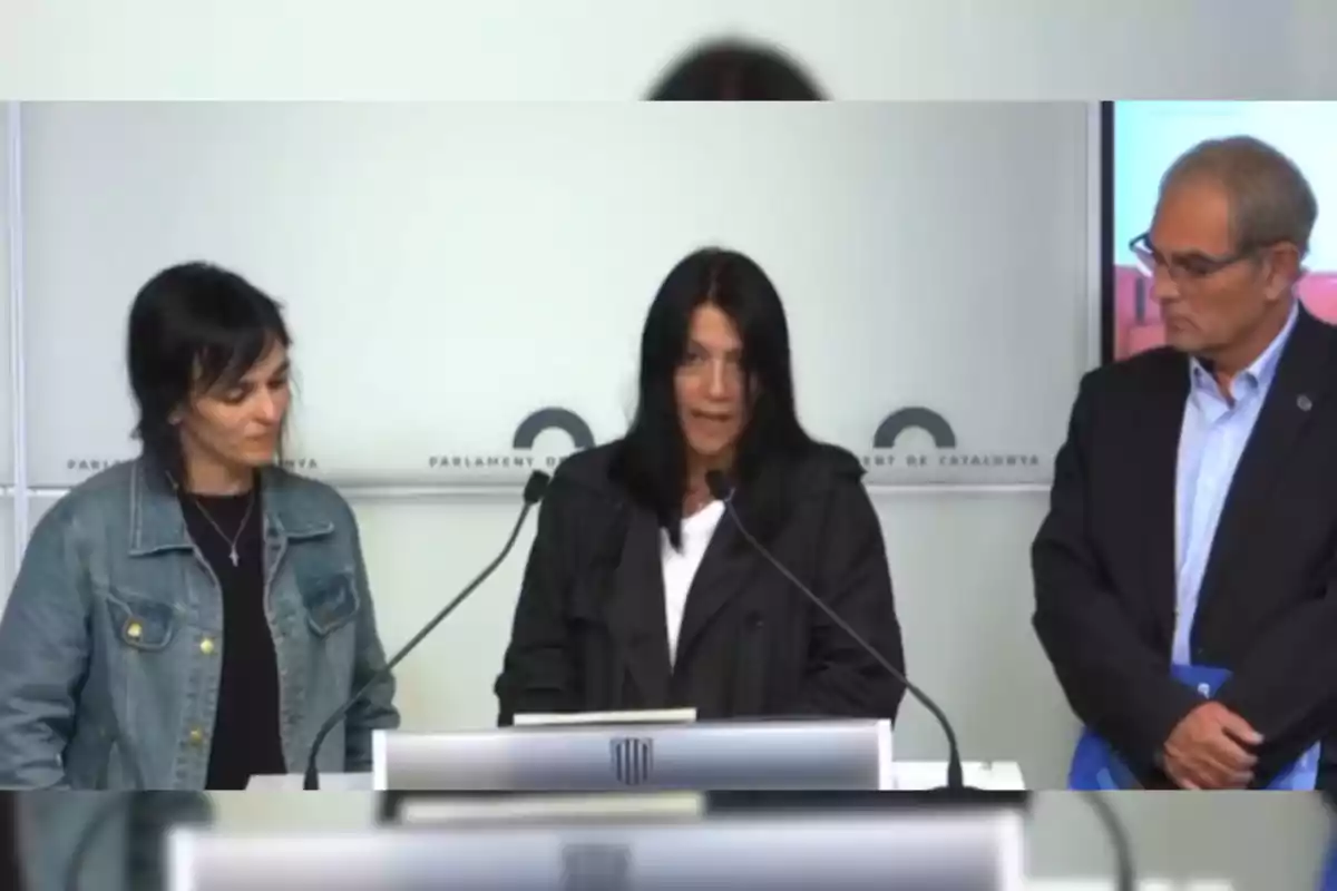 Tres personas están de pie frente a un podio en una conferencia de prensa en el Parlament de Catalunya.