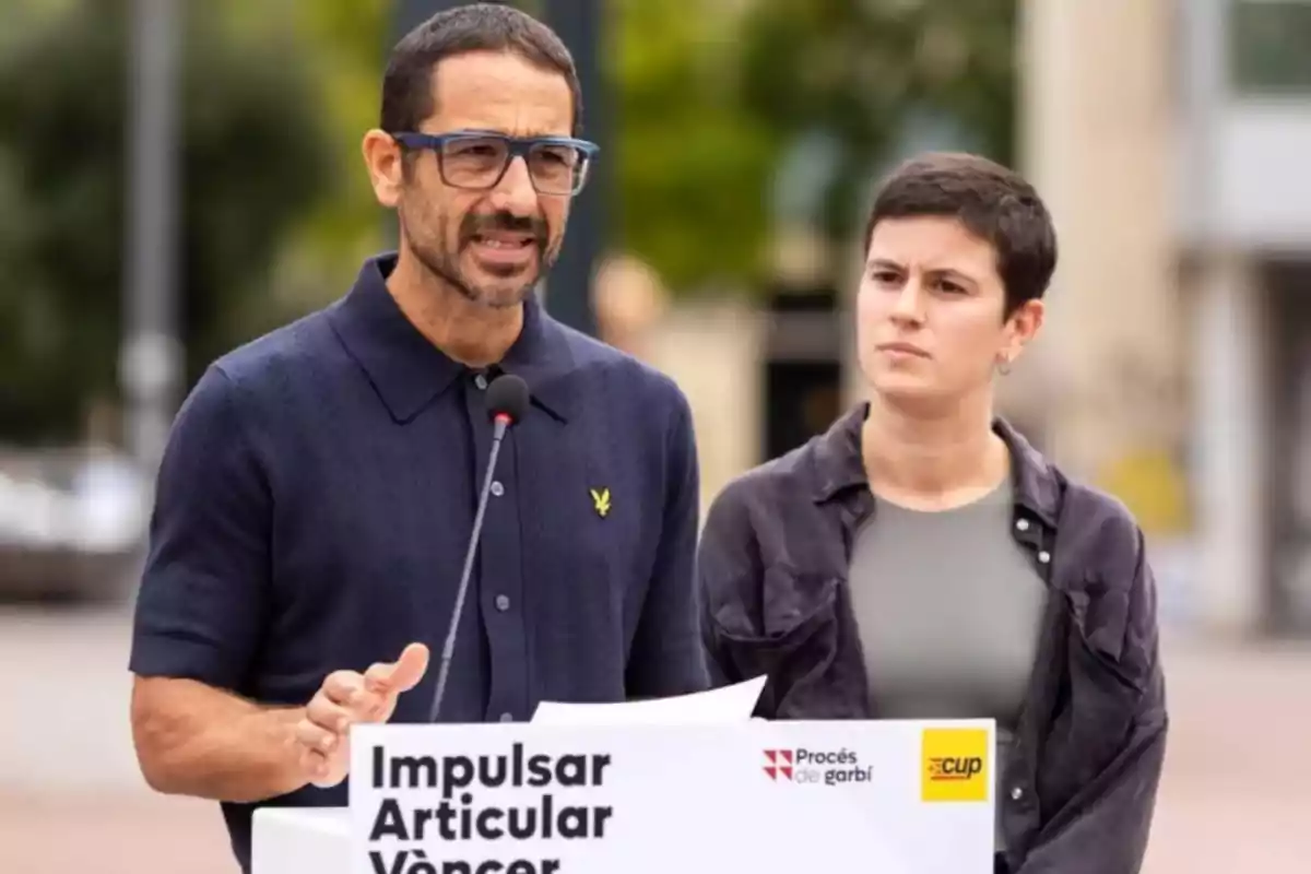 Dos personas en una conferencia de prensa al aire libre con un micrófono y un cartel que dice "Impulsar Articular Vèncer".