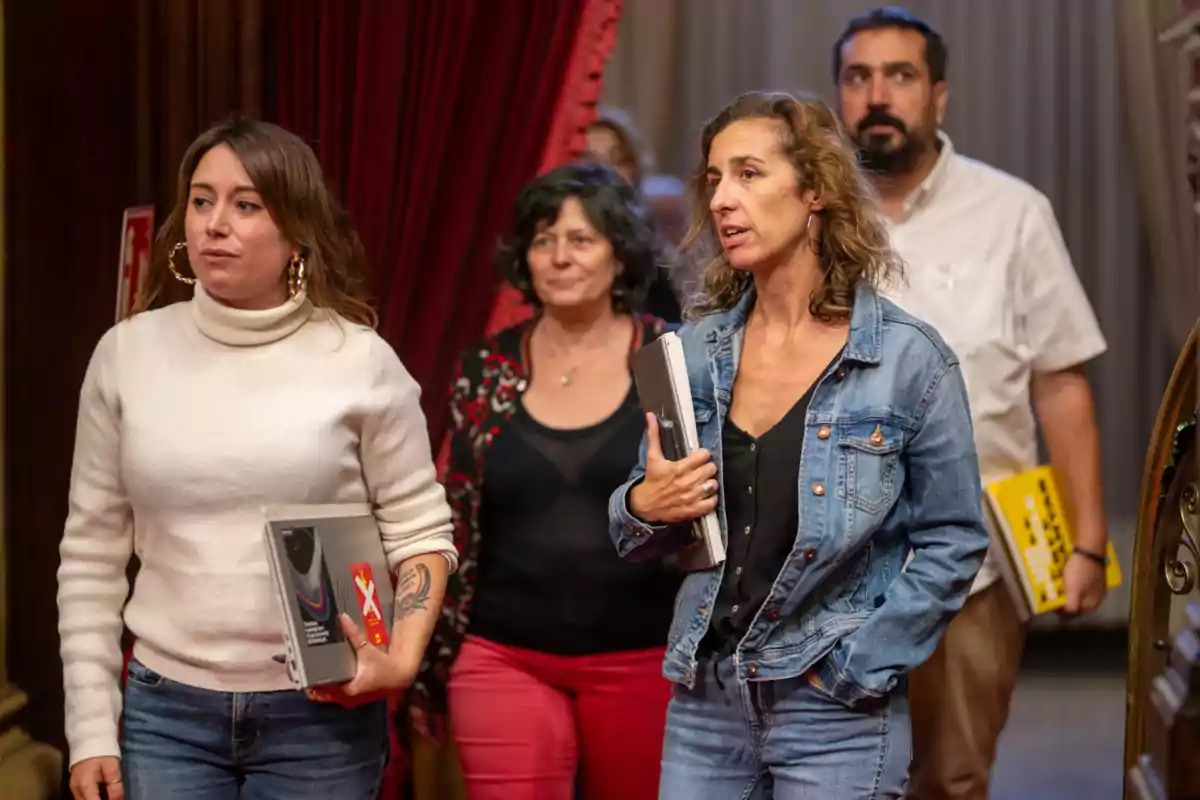 Un grupo de personas camina por un pasillo con cortinas rojas, llevando carpetas y libros.