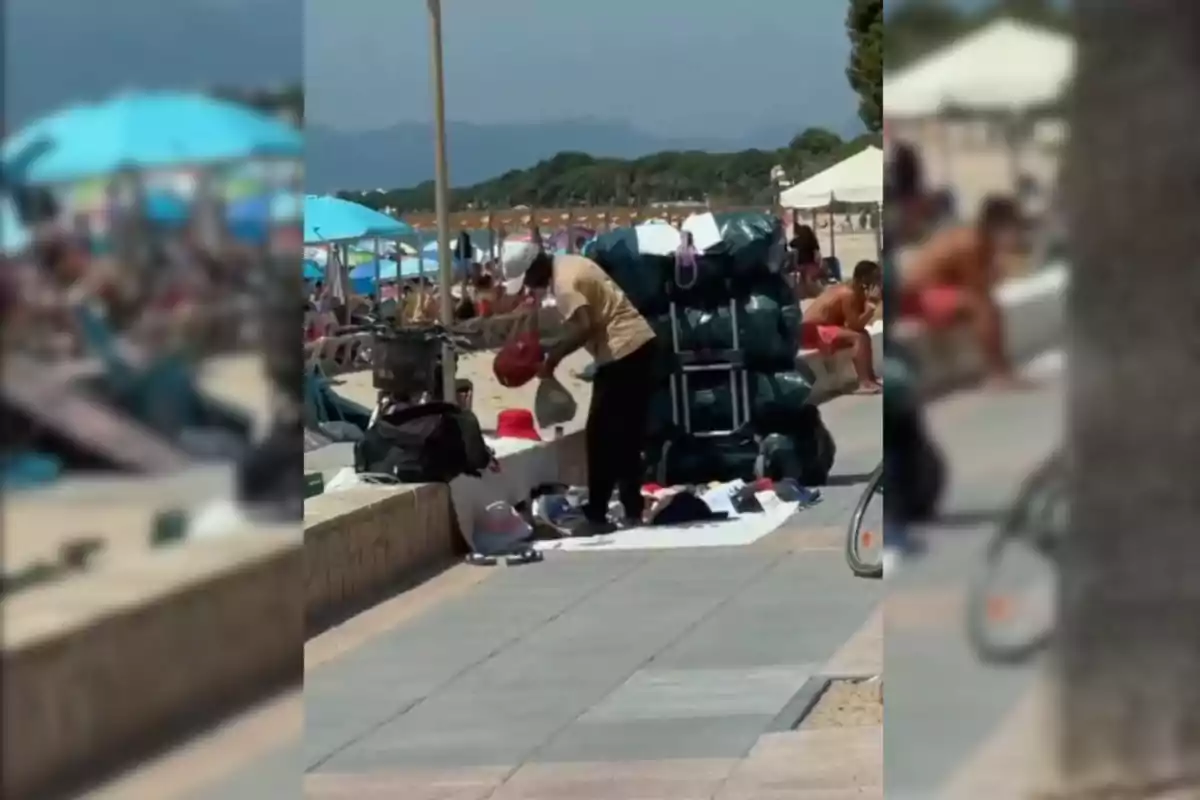 Un mantero organizando mercancía en una playa concurrida con sombrillas y turistas al fondo.