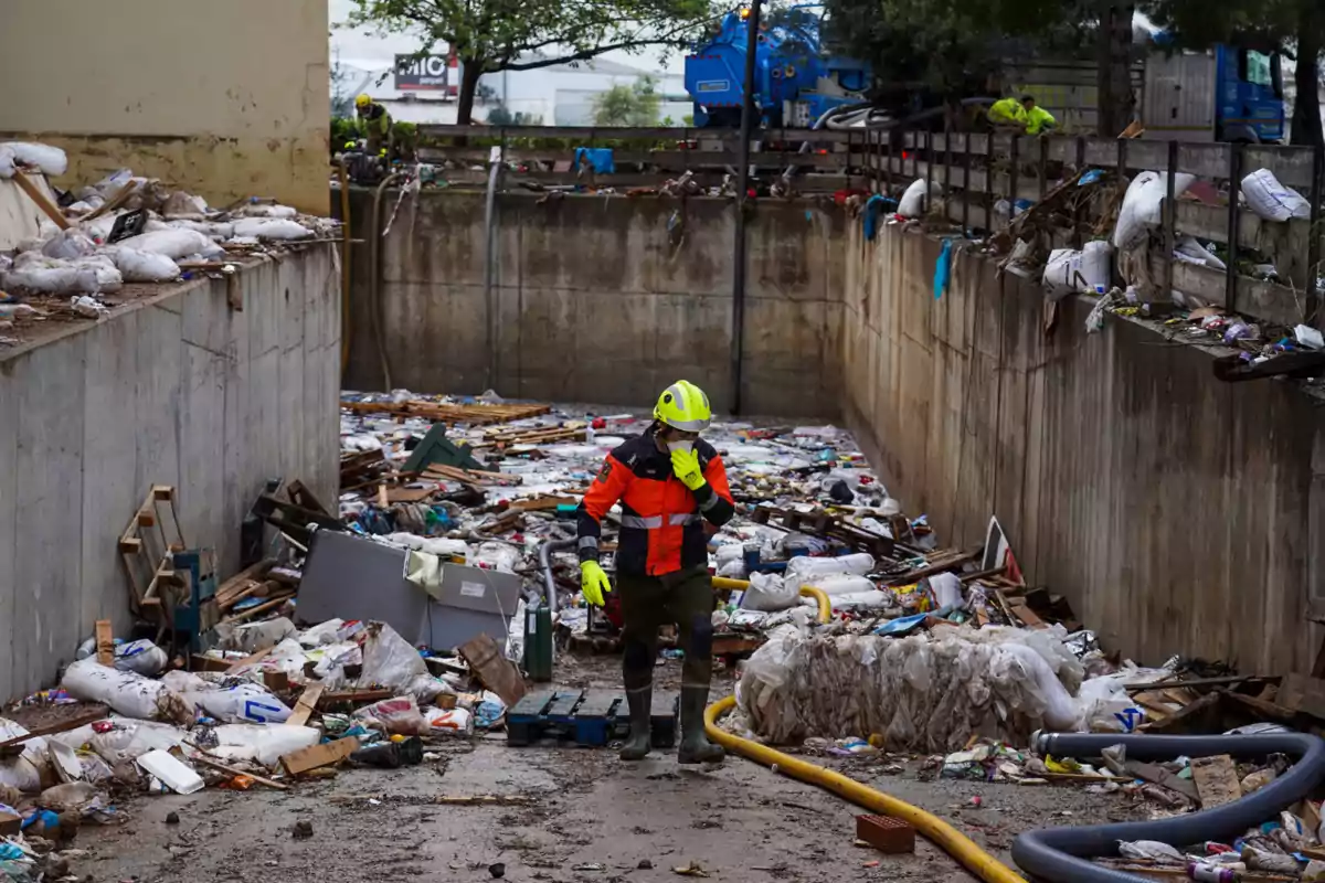 Un trabajador con casco y chaleco de seguridad camina entre escombros y basura en un área cercada de concreto.