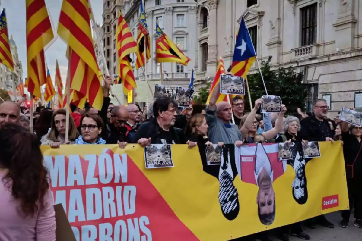 Un grupo de personas participa en una manifestación en la calle, sosteniendo una pancarta amarilla y roja con texto y varias banderas con franjas amarillas y rojas.