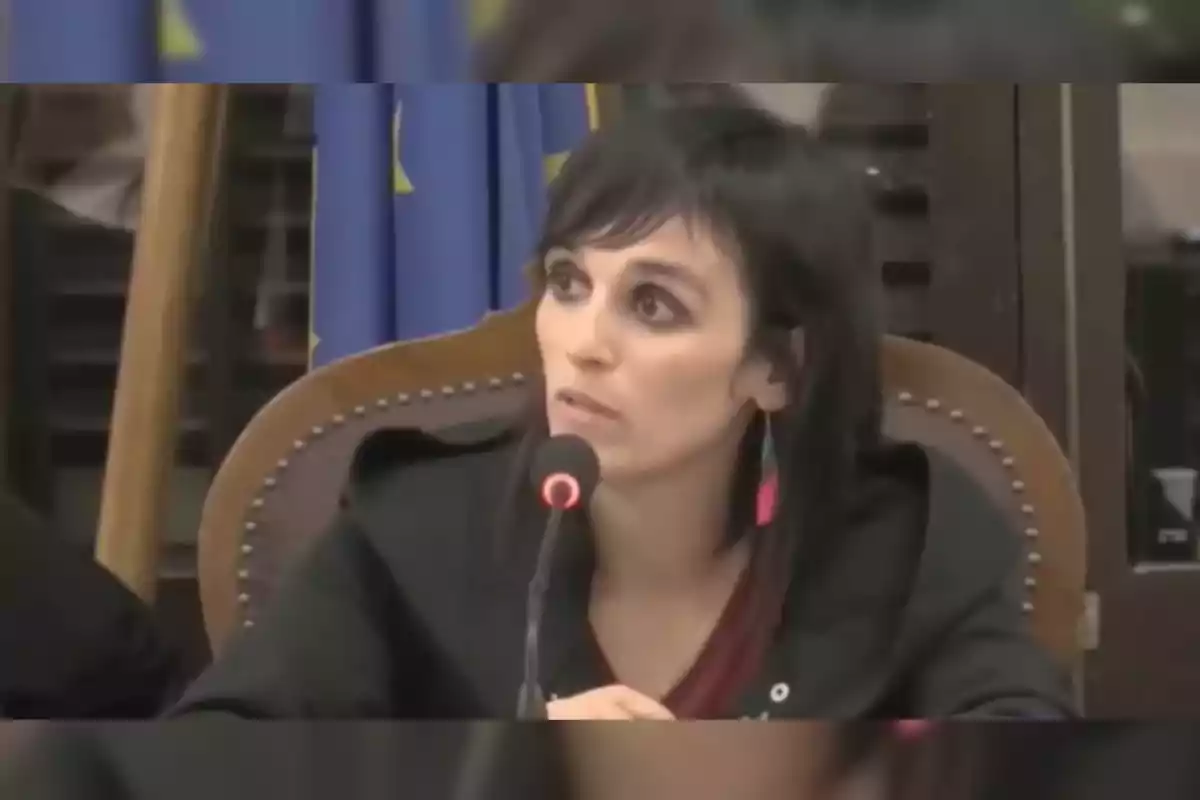 Sílvia Orriols hablando en un micrófono durante una conferencia con una bandera de la Unión Europea de fondo.