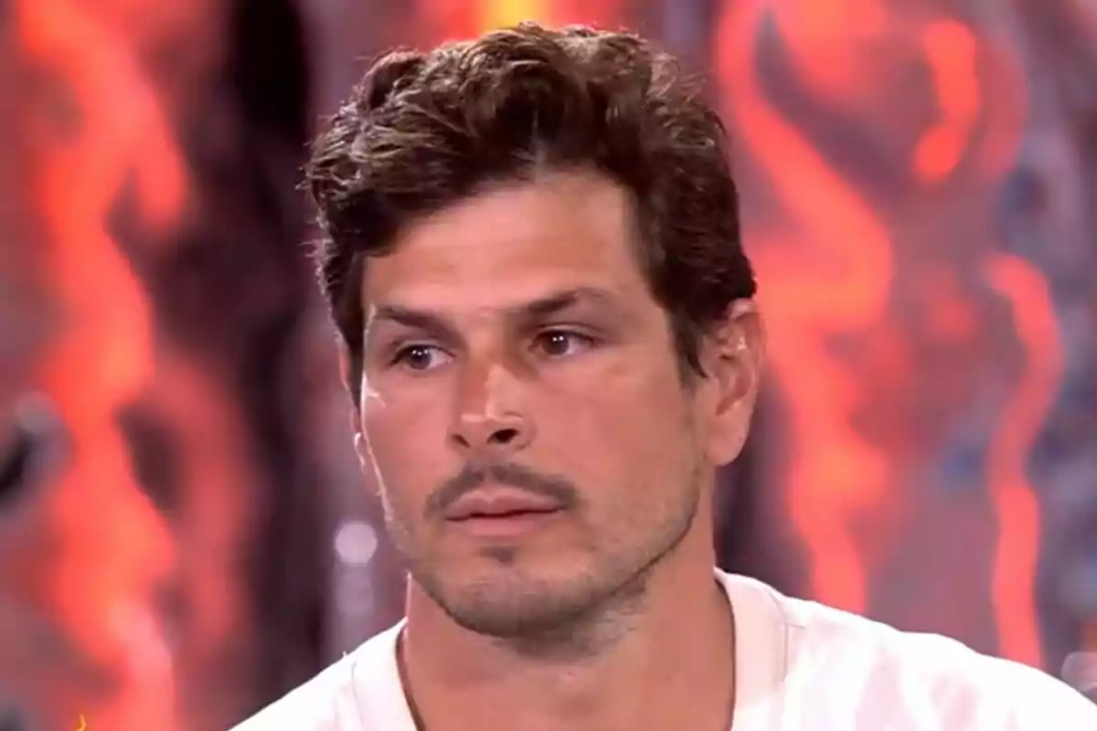 Captura de Alejandro Nieto con cabello corto y barba ligera, vistiendo una camiseta blanca, con un fondo desenfocado de colores cálidos en el plató de Supervivientes