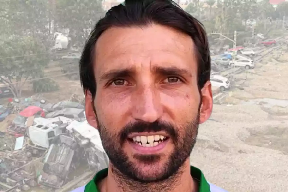 Un hombre con barba y cabello oscuro frente a un fondo de autos dañados y árboles.