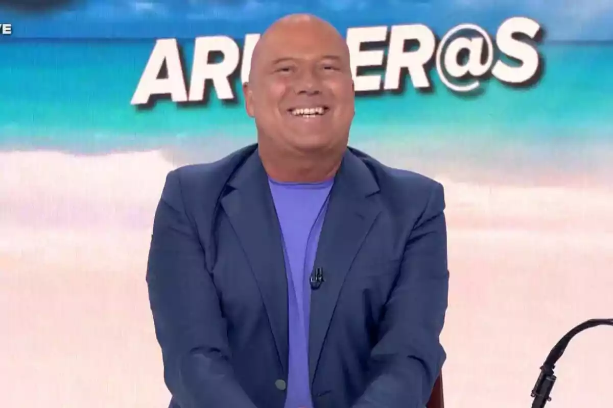 Captura de Alfonso Arús sonriendo mientras está sentado, vistiendo un traje azul con una camiseta morada, con un fondo que muestra la palabra "ARUSER@S" y una imagen de playa.